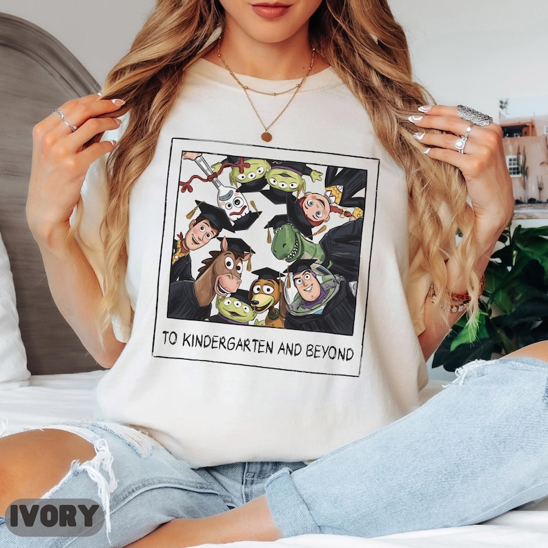 May include: A cream-colored t-shirt featuring a graphic print of Toy Story characters wearing graduation caps inside a polaroid frame. The text below the characters reads TO KINDERGARTEN AND BEYOND. The shirt is paired with light blue denim jeans.