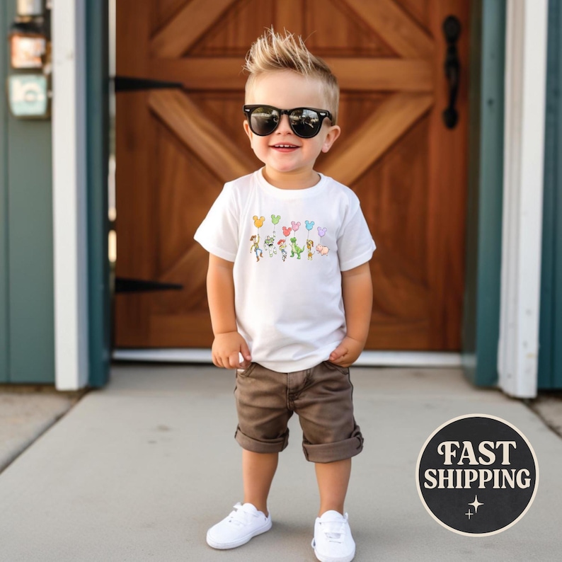 K&ouml;nnte beinhalten: Ein Kind tr&auml;gt ein wei&szlig;es T-Shirt mit einem bunten Cartoon-Design, braune Shorts, wei&szlig;e Turnschuhe und eine schwarze Sonnenbrille. Das Shirt zeigt verschiedene Cartoon-Figuren und Ballons. Das Kind steht vor einer Holzt&uuml;r.