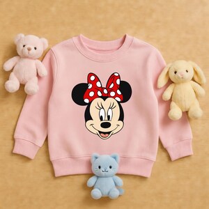 Może przedstawiać: Jasnor&oacute;żowa bluza z nadrukiem Myszki Minnie z czerwoną kokardą w białe grochy. Na zdjęciu r&oacute;żowy miś, ż&oacute;łty kr&oacute;lik i niebieski pluszowy kot.