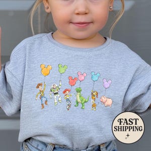 K&ouml;nnte beinhalten: Ein hellgraues Sweatshirt mit einer Aquarellillustration von Toy Story-Charakteren, die Mickey Mouse-f&ouml;rmige Ballons in verschiedenen Farben halten. Zu den Charakteren geh&ouml;ren Woody, Buzz Lightyear und Rex. Eine kreisf&ouml;rmige "FAST SHIPPING"-Grafik befindet sich unten rechts.