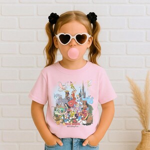 May include: A light pink t-shirt featuring a colorful graphic of various Disney characters, including Dumbo, Peter Pan, and Mickey Mouse. The text "Magic Kingdom, Walt Disney World" is printed below the characters. The shirt is worn by a person wearing heart-shaped sunglasses.