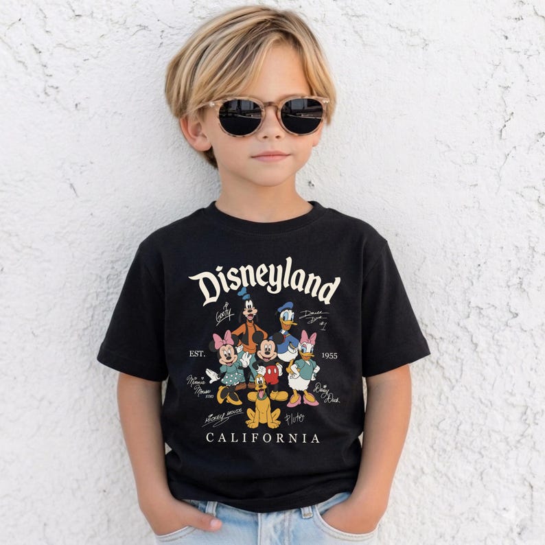 May include: A black t-shirt featuring the word "Disneyland" in white script, along with cartoon characters like Mickey Mouse, Minnie Mouse, and Donald Duck. The shirt also has the text "EST. 1955 CALIFORNIA". The child is wearing sunglasses.