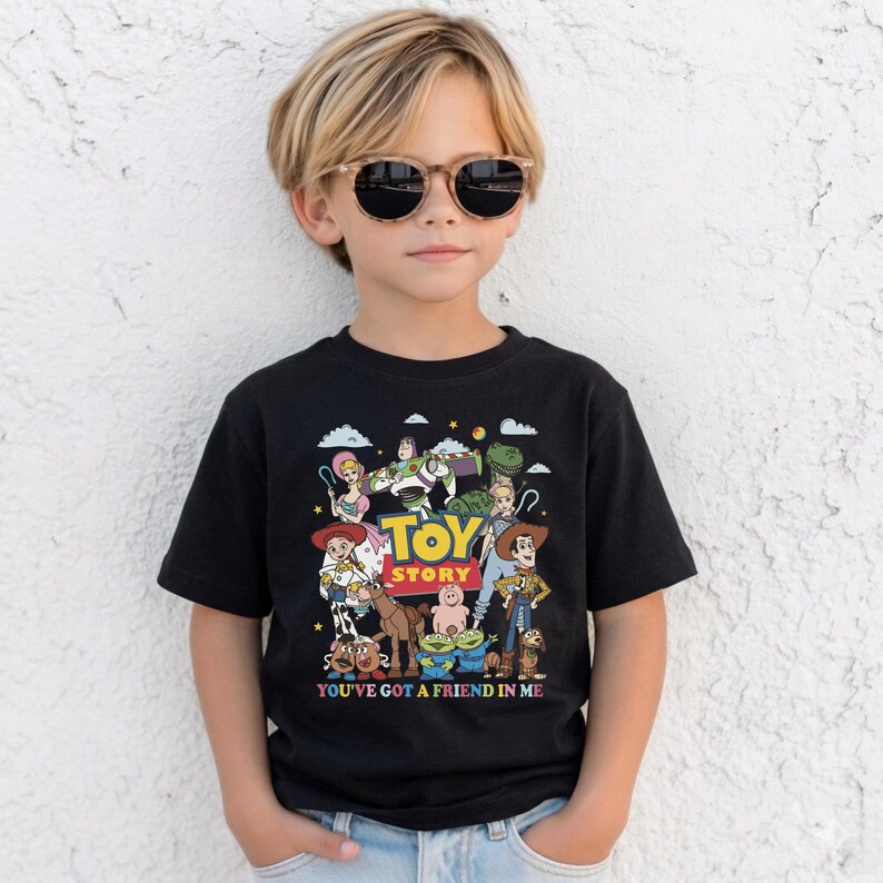Op de afbeelding: Zwart T-shirt met een kleurrijke Toy Story-afbeelding, met Woody, Buzz Lightyear en andere personages. De tekst "TOY STORY" staat in het midden, met "YOU'VE GOT A FRIEND IN ME" eronder. Het shirt wordt door een kind gedragen.