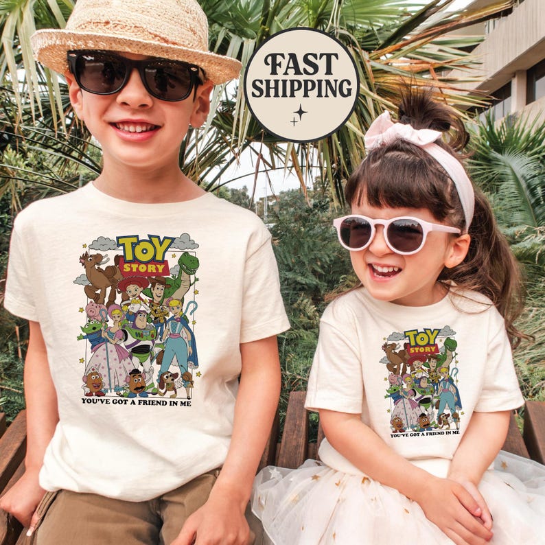 Puede incluir: Dos ni&ntilde;os con camisetas color crema con un gr&aacute;fico colorido de personajes de Toy Story. Las camisetas tienen el texto "TOY STORY" y "YOU'VE GOT A FRIEND IN ME". El ni&ntilde;o lleva gafas de sol y un sombrero de paja. La ni&ntilde;a lleva gafas de sol y un lazo rosa.