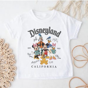 May include: White t-shirt featuring a Disneyland graphic with cartoon characters like Mickey Mouse, Minnie Mouse, Goofy, Donald Duck, and Daisy Duck. The graphic includes the text "Disneyland California" and the year "1955".
