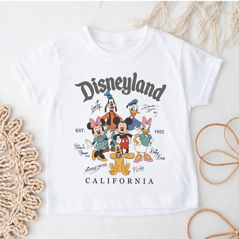 May include: White t-shirt featuring a Disneyland graphic with cartoon characters like Mickey Mouse, Minnie Mouse, Goofy, Donald Duck, and Daisy Duck. The graphic includes the text "Disneyland California" and the year "1955".
