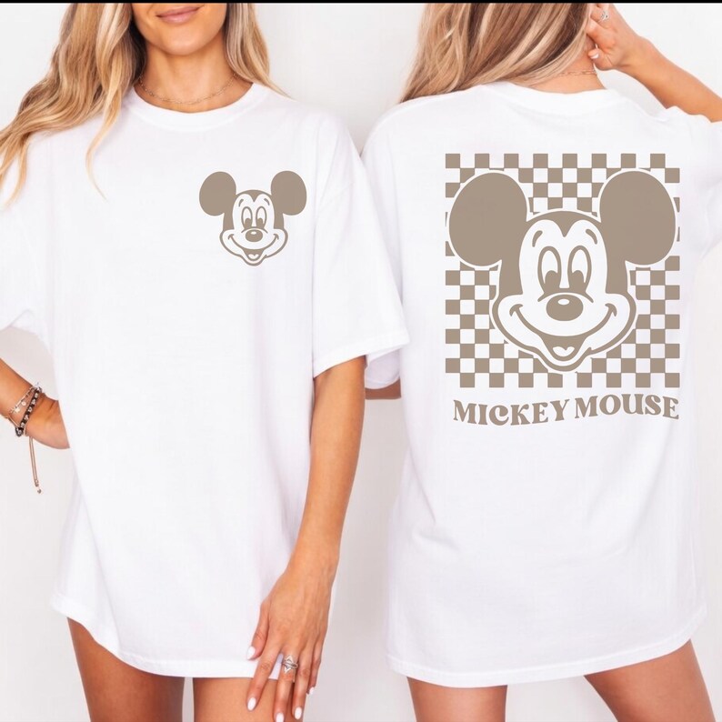 May include: White t-shirt featuring a taupe Mickey Mouse graphic on the front and back. The back design includes a checkered pattern and the words "Mickey Mouse". The shirt has short sleeves and a relaxed fit.