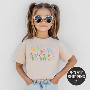 K&ouml;nnte beinhalten: Beiges T-Shirt mit einer Illustration von Zeichentrickfiguren, die Mickey Mouse-f&ouml;rmige Ballons halten. Zu den Figuren geh&ouml;ren Woody, Buzz Lightyear, Jessie, Rex und Hamm. Das T-Shirt wird von einer jungen Person getragen, die herzf&ouml;rmige Sonnenbrillen tr&auml;gt.