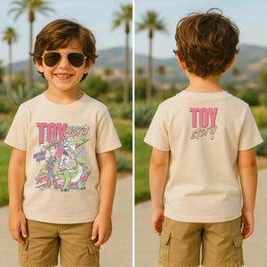 May include: A beige t-shirt featuring the text "Toy Story" in pink and teal, with cartoon characters. The shirt is worn by a child wearing sunglasses and khaki shorts. The back of the shirt also has the text "Toy Story" in pink.