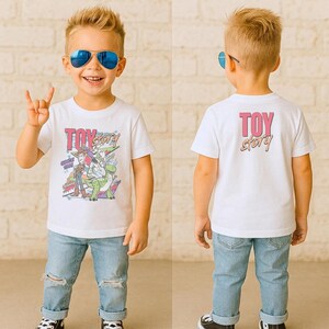 May include: A white t-shirt featuring the text "TOY STORY" in pink and a graphic of characters from the movie. The shirt is worn by a young child with blue sunglasses and ripped blue jeans.