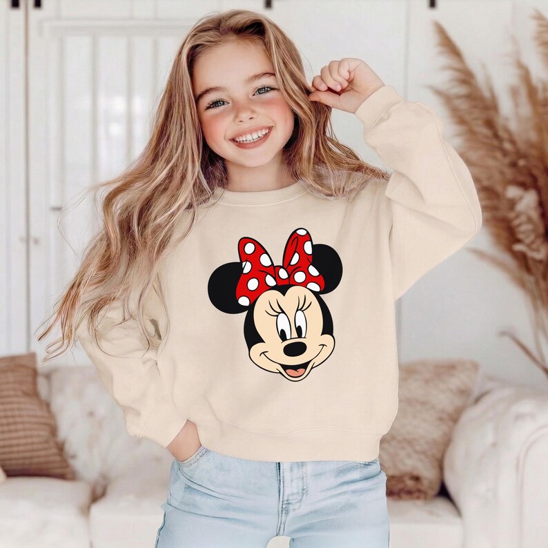 Może przedstawiać: Kremowa bluza z nadrukiem Myszki Minnie. Grafika przedstawia twarz Myszki Minnie z czerwoną kokardą w białe grochy. Bluza ma długie rękawy i okrągły dekolt. Styl jest swobodny i odpowiedni dla dzieci.