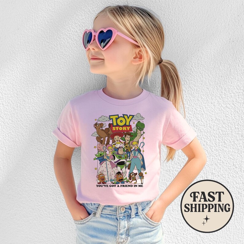 Puede incluir: Una camiseta rosa claro con un gr&aacute;fico colorido de personajes de la pel&iacute;cula animada "Toy Story". La camiseta tiene las mangas enrolladas y el texto "TOY STORY" y "YOU'VE GOT A FRIEND IN ME". Una persona joven lleva la camiseta y gafas de sol en forma de coraz&oacute;n.