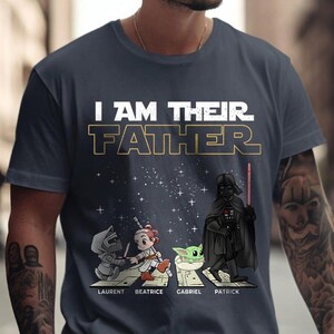 May include: Navy blue t-shirt with the text "I AM THEIR FATHER" in white and gold. The shirt features cartoon illustrations of Star Wars characters, including Darth Vader, and the names Laurent, Beatrice, Gabriel, and Patrick.