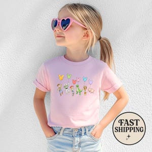 K&ouml;nnte beinhalten: Hellrosa T-Shirt mit einem Cartoon-Design von Figuren, die Mickey Mouse-f&ouml;rmige Ballons halten. Das T-Shirt wird von einer jungen Person getragen, die eine rosa herzf&ouml;rmige Sonnenbrille tr&auml;gt.