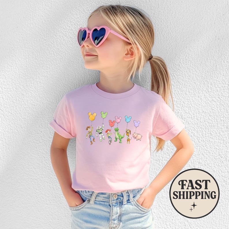 K&ouml;nnte beinhalten: Hellrosa T-Shirt mit einem Cartoon-Design von Figuren, die Mickey Mouse-f&ouml;rmige Ballons halten. Das T-Shirt wird von einer jungen Person getragen, die eine rosa herzf&ouml;rmige Sonnenbrille tr&auml;gt.