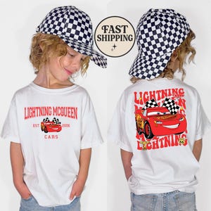 Puede incluir: Camiseta blanca con gr&aacute;ficos de Lightning McQueen de la pel&iacute;cula Cars. La parte delantera muestra "Lightning McQueen" y un gr&aacute;fico del coche. La parte trasera presenta un gr&aacute;fico m&aacute;s grande del coche con el texto "Lightning" y "Cars". Tambi&eacute;n se ve una gorra a cuadros.