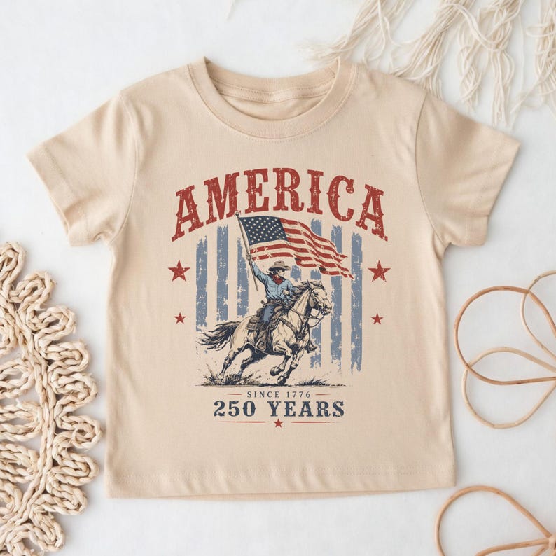 May include: A beige short-sleeve t-shirt featuring a graphic of a person riding a horse while holding an American flag. The text reads AMERICA, SINCE 1776, and 250 YEARS. The design includes red stars and a distressed blue and white striped background.