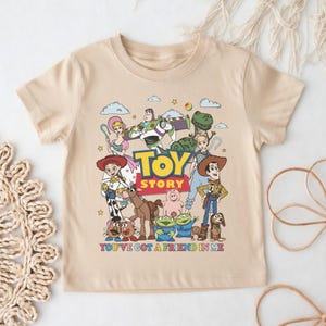 Op de afbeelding: Een beige t-shirt met een kleurrijke afbeelding van personages uit de film "Toy Story". Het ontwerp bevat Woody, Buzz Lightyear en andere personages, met de woorden "Toy Story" en "You've Got a Friend in Me".