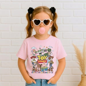 Op de afbeelding: Een lichtroze t-shirt met een kleurrijke afbeelding van personages uit de animatiefilm "Toy Story". Op het shirt staan de woorden "Toy Story" en "You've Got a Friend in Me". Het shirt wordt door een jong meisje gedragen.
