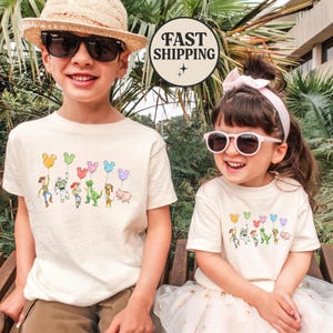 K&ouml;nnte beinhalten: Zwei Kinder tragen cremefarbene T-Shirts mit einer bunten Grafik von Zeichentrickfiguren, die ballonf&ouml;rmige Ballons halten. Die Grafik enth&auml;lt Charaktere wie Woody, Buzz Lightyear und Rex. Die Shirts sind kurz&auml;rmlig.