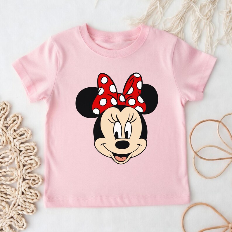 Może przedstawiać: Jasnor&oacute;żowa koszulka dla malucha z grafiką Myszki Minnie. Grafika przedstawia twarz Myszki Minnie z czerwoną kokardą w białe grochy. Koszulka ma kr&oacute;tkie rękawy i okrągły dekolt.