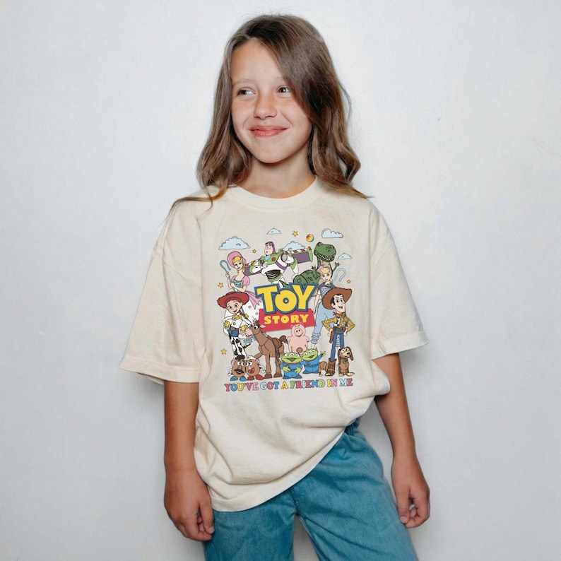 Op de afbeelding: Cr&egrave;mekleurig T-shirt met een kleurrijke afbeelding van personages uit de animatiefilm "Toy Story". Het ontwerp bevat Woody, Buzz Lightyear en andere personages, met de tekst "TOY STORY" en "YOU'VE GOT A FRIEND IN ME".
