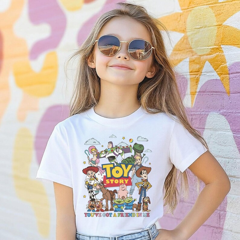 Op de afbeelding: Een wit T-shirt met een kleurrijke afbeelding van personages uit de animatiefilm "Toy Story". Het ontwerp bevat Woody, Buzz Lightyear en andere personages, met de tekst "TOY STORY" en "YOU'VE GOT A FRIEND IN ME."