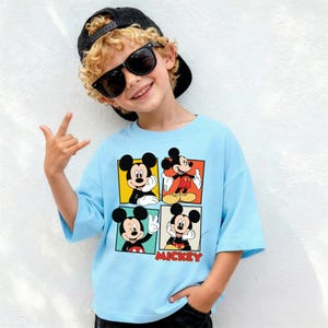 May include: A light blue t-shirt with a four-panel Mickey Mouse design. The word "Mickey" is printed in red. The shirt is worn by a person wearing sunglasses and a black cap.