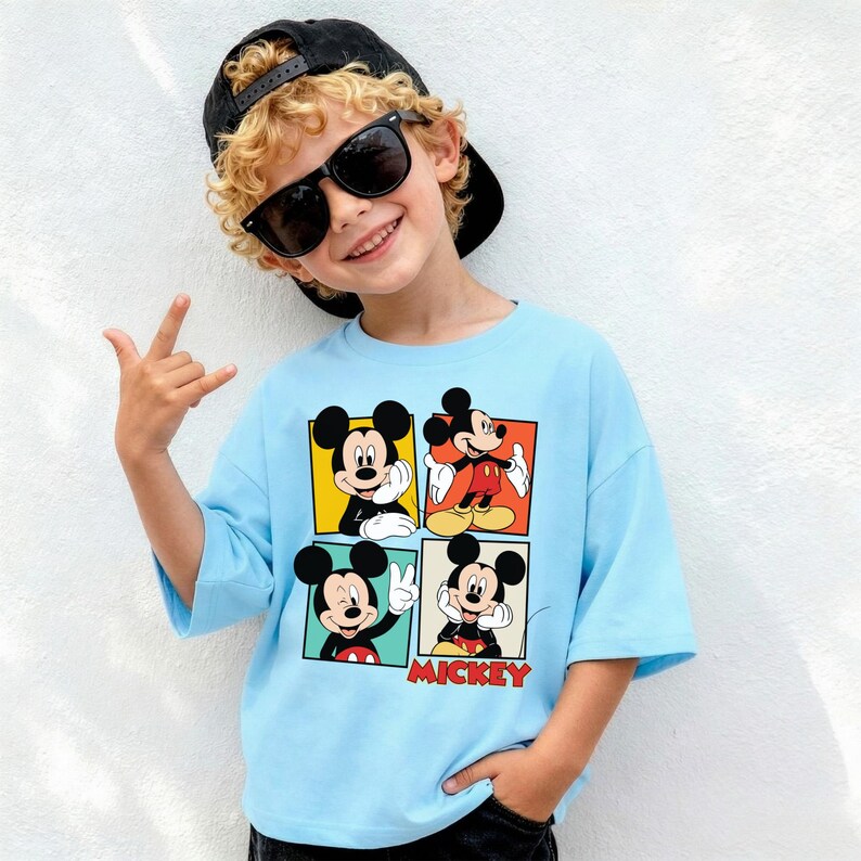 May include: A light blue t-shirt with a four-panel Mickey Mouse design. The word "Mickey" is printed in red. The shirt is worn by a person wearing sunglasses and a black cap.