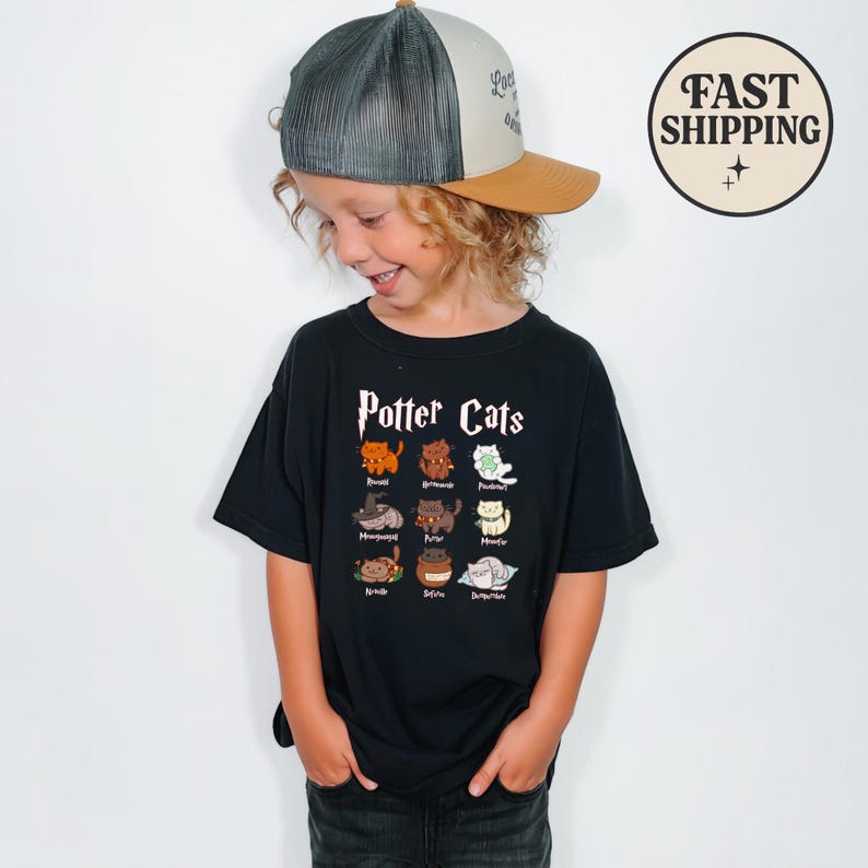 May include: A black t-shirt featuring the words "Potter Cats" and illustrations of various cartoon cats. The child is wearing a trucker hat with a gray mesh back and a tan brim. The hat has the words "Love is all" on the front.