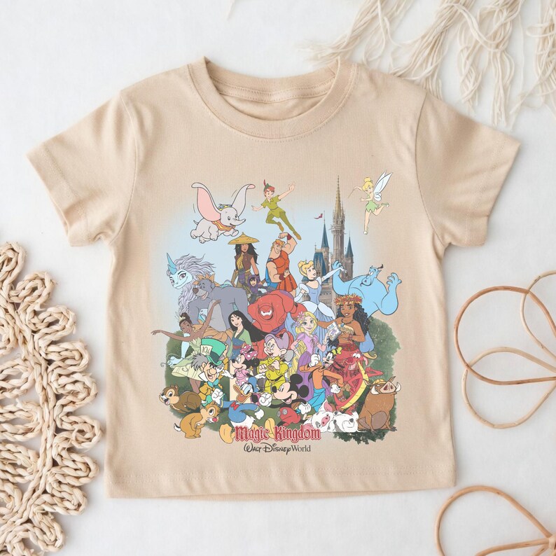 May include: Beige short-sleeved t-shirt featuring a colorful graphic of Disney characters, including Mickey Mouse, Dumbo, and Peter Pan. The text "Magic Kingdom Walt Disney World" is printed at the bottom of the design.