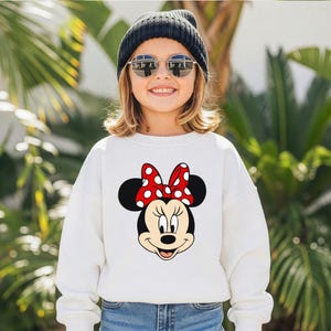 Może przedstawiać: Biała bluza z grafiką Myszki Minnie. Grafika przedstawia twarz Myszki Minnie z czerwoną kokardą w białe grochy. Bluza jest noszona przez osobę noszącą czarną czapkę i okulary przeciwsłoneczne.