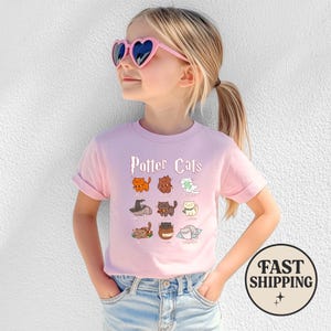 May include: A light pink t-shirt featuring the text "Potter Cats" above a grid of cartoon cat illustrations. The cats are in various colors and poses, with some wearing hats and accessories. The shirt is worn by a young girl wearing pink heart-shaped sunglasses.