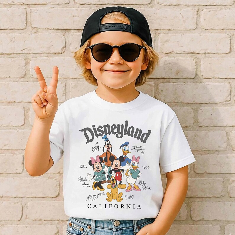 May include: A white t-shirt featuring the Disneyland logo and cartoon characters like Mickey Mouse, Minnie Mouse, and Donald Duck. The shirt has the text "EST. 1955 CALIFORNIA". The person is wearing a black cap and sunglasses.