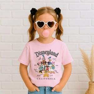 May include: A light pink t-shirt with the word "Disneyland" in a script font. The shirt features cartoon characters including Mickey Mouse, Minnie Mouse, Donald Duck, and Goofy. The text "CALIFORNIA" is printed below the characters.