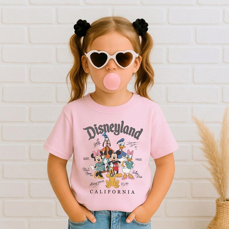 May include: A light pink t-shirt with the word "Disneyland" in a script font. The shirt features cartoon characters including Mickey Mouse, Minnie Mouse, Donald Duck, and Goofy. The text "CALIFORNIA" is printed below the characters.