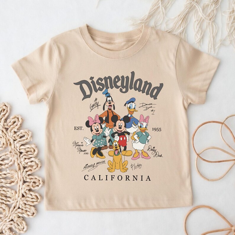 May include: A beige t-shirt featuring the word "Disneyland" in a stylized font, with cartoon characters including Mickey Mouse, Minnie Mouse, Goofy, Donald Duck, Daisy Duck, and Pluto. The text "CALIFORNIA" is at the bottom.