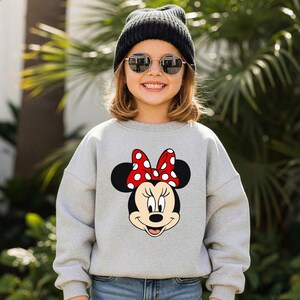 Może przedstawiać: Szara bluza z nadrukiem Myszki Minnie. Grafika przedstawia twarz Myszki Minnie z czerwoną kokardą w białe grochy. Osoba na zdjęciu ma na sobie czarną czapkę i okrągłe okulary przeciwsłoneczne.