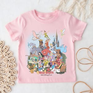 May include: A light pink t-shirt featuring a colorful graphic of various Disney characters, including Mickey Mouse, Dumbo, and Peter Pan. The words "Magic Kingdom" and "Walt Disney World" are printed at the bottom of the design.