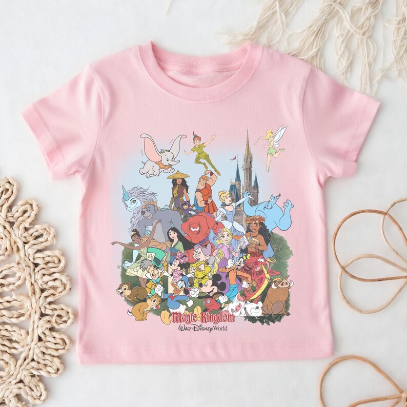 May include: A light pink t-shirt featuring a colorful graphic of various Disney characters, including Mickey Mouse, Dumbo, and Peter Pan. The words "Magic Kingdom" and "Walt Disney World" are printed at the bottom of the design.