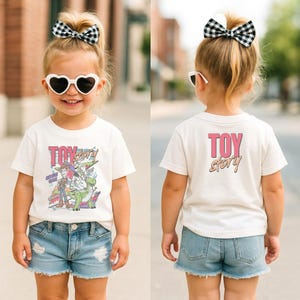 May include: A child wearing a white t-shirt with a "Toy Story" graphic on the front and back. The front graphic features characters from the movie. The child is also wearing denim shorts, heart-shaped sunglasses, and a black and white checkered bow.