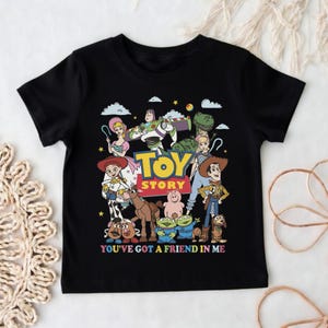 Op de afbeelding: Zwart T-shirt met een kleurrijke afbeelding van personages uit de animatiefilm "Toy Story". Het ontwerp bevat Woody, Buzz Lightyear en andere personages, met de tekst "TOY STORY" en "YOU'VE GOT A FRIEND IN ME."