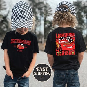 Puede incluir: Camiseta negra con Lightning McQueen de la pel&iacute;cula Cars. El frente muestra "Lightning McQueen" y "Cars". La parte trasera muestra a McQueen con banderas a cuadros y el texto "Lightning". El ni&ntilde;o lleva una gorra a cuadros en blanco y negro.