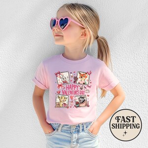 May include: A pink t-shirt featuring a graphic with four framed cartoon characters and the text "Happy Valentine's Day." The shirt is worn by a young girl wearing pink heart-shaped sunglasses.