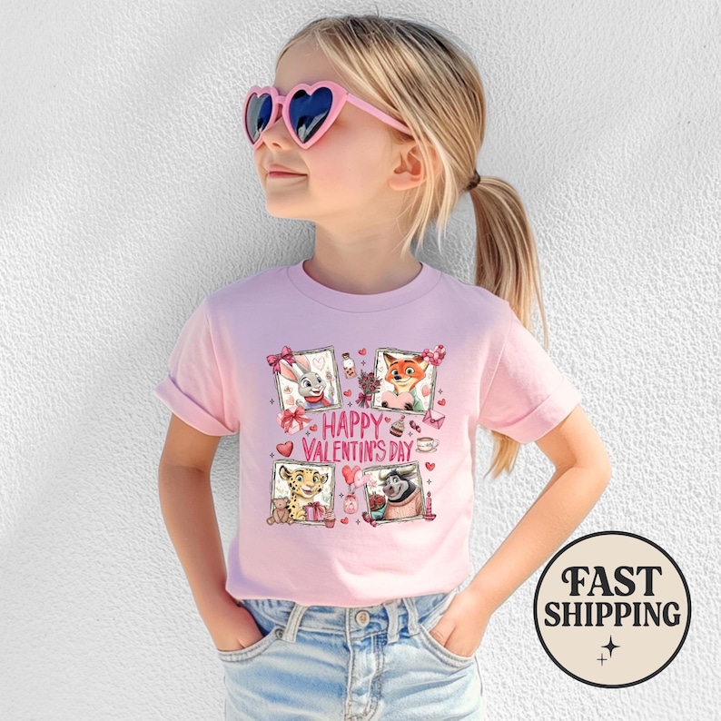 May include: A pink t-shirt featuring a graphic with four framed cartoon characters and the text "Happy Valentine's Day." The shirt is worn by a young girl wearing pink heart-shaped sunglasses.