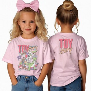 May include: Two young girls wearing light pink t-shirts with a "Toy Story" graphic. The front of the shirt features Woody, Buzz Lightyear, and Rex. The back of the shirt has the words "Toy Story". The girls are wearing blue jeans and a pink bow.