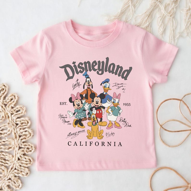 May include: A light pink t-shirt featuring the word "Disneyland" in a stylized font, along with cartoon characters like Mickey Mouse, Minnie Mouse, and Donald Duck. The text "CALIFORNIA" is printed at the bottom.