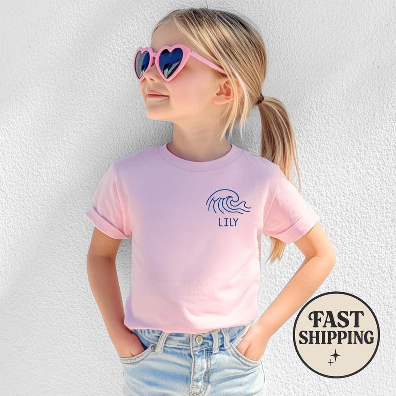 Op de afbeelding: Een lichtroze t-shirt met opgerolde mouwen. Het shirt heeft een marineblauwe golfafbeelding en de naam "LILY" eronder gedrukt. Het kind draagt hartvormige zonnebrillen en een blauwe jeans.