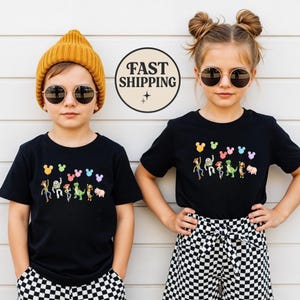 K&ouml;nnte beinhalten: Zwei Kinder tragen schwarze T-Shirts mit einer bunten Cartoon-Grafik von Figuren und Luftballons. Die Shirts haben ein Design im Mickey-Mouse-Stil &uuml;ber den Figuren. Ein Kind tr&auml;gt eine gelbe M&uuml;tze und das andere eine Sonnenbrille. Der Hintergrund ist eine wei&szlig;e Wand.