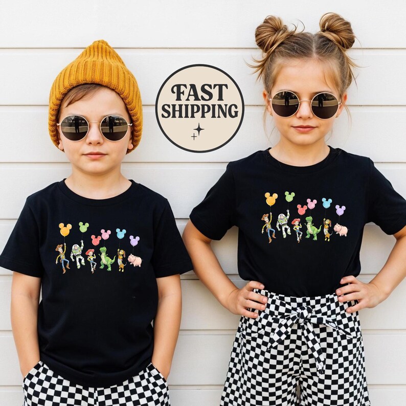 K&ouml;nnte beinhalten: Zwei Kinder tragen schwarze T-Shirts mit einer bunten Cartoon-Grafik von Figuren und Luftballons. Die Shirts haben ein Design im Mickey-Mouse-Stil &uuml;ber den Figuren. Ein Kind tr&auml;gt eine gelbe M&uuml;tze und das andere eine Sonnenbrille. Der Hintergrund ist eine wei&szlig;e Wand.